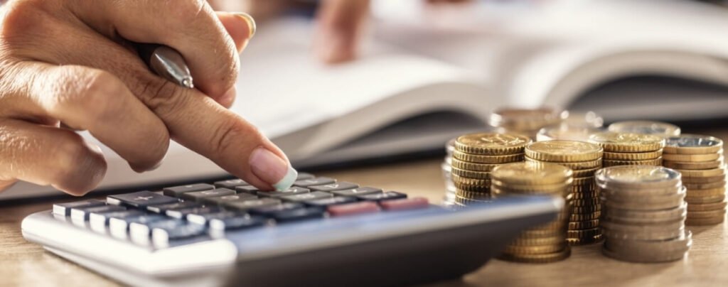 A close-up of a person's hand using a calculator next to stacks of coins, symbolizing the process of calculating predial (property tax) or financial investment returns.