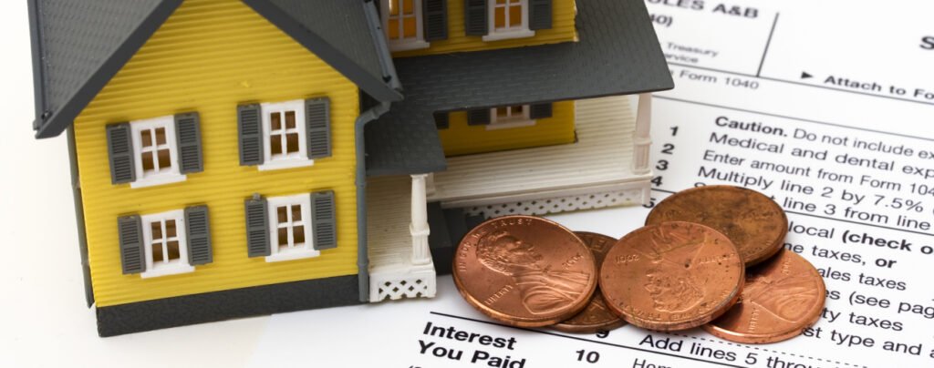 A miniature yellow and grey house model and a pile of copper pennies resting on a tax form (Form 1040), symbolizing property tax and home finance.
