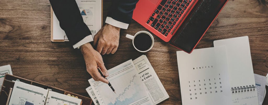 A high-angle, top-down view of a dark wooden desk where a person in a dark business suit and orange-rimmed glasses is pointing a pen at a line graph on a printed document. The desk is cluttered with professional items, including a silver laptop, a cup of black coffee, a leather-bound planner, a "Tax Refund Form," a calendar, and a smartphone. Two other sets of hands are visible at the bottom of the frame, suggesting a collaborative business meeting or legal consultation.