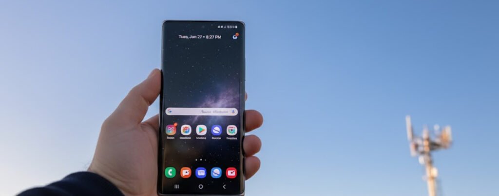 A first-person perspective of a hand holding an older, white smartphone against a clear blue sky. The phone’s screen is mostly blank white, displaying only a green cellular signal strength icon in the top left corner. In the blurred background, a cell tower stands in the distance.