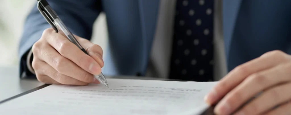 A person wearing a medium-blue suit and a navy polka-dot tie holds a pen and signs a paper on a black clipboard.